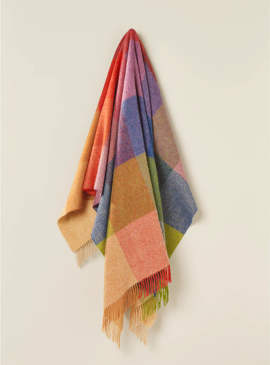 A multicolored wool throw with a geometric pattern, featuring shades of yellow, orange, blue, green, red, and purple, hanging on a wall.